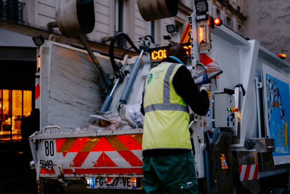 A waste collection worker wearing a high-visibility yellow vest and dark clothing is operating an industrial rubbish collection vehicle on a city street during dusk or early evening. The vehicle is equipped with hydraulic arms and sorting mechanisms, with its rear open to reveal a large interior compartment filled with collected waste materials. The truck's exterior is white with red reflective safety markings and emergency lights, which are illuminated. The worker is standing on the pavement, managing controls on the side of the vehicle. In the background, there are urban buildings with lit windows, indicating a busy area that may experience congestion or restricted access issues, relevant to specialist waste removal services for private or alternative waste handling on busy roads or narrow streets. The lighting creates a contrast between the brightly lit truck and the dimmer surroundings, emphasizing the operational nature of the scene and supporting the context of independent rubbish collection for road access challenges in areas like Kensington.
