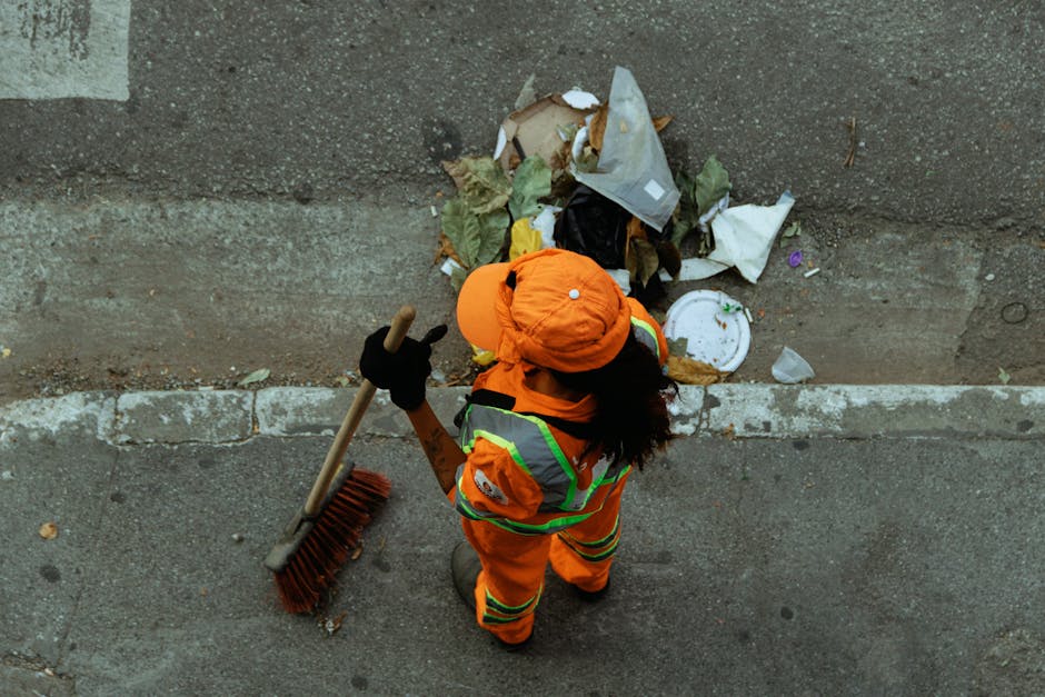 An individual dressed in high-visibility orange reflective work clothing, including a matching hat and gloves, is standing on a paved sidewalk next to a concrete wall with a slightly raised edge. The person is holding a long-handled broom with a wooden handle and orange bristles, positioned to sweep debris. In front of them, scattered along the base of the wall, there is a pile of mixed waste materials, including crumpled paper, plastic packaging, a white disposable plate, black and white plastic bags, and various organic waste such as leaves and food wrappers. The debris appears loosely piled, with some items extending onto the sidewalk surface, which shows small cracks and embedded dirt. The natural daylight illuminates the scene evenly, emphasizing the contrast between the bright safety gear and the dull grey tones of the pavement and wall. This image illustrates a scene of on-site waste clearance, possibly performed by Waste Removal Kensington, highlighting typical challenges encountered during private rubbish collection on urban roads around Earls Court, Kensington.