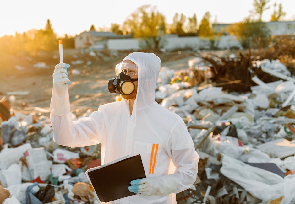 A person dressed in full protective gear, including a white coverall, gloves, a face mask with a respirator, and goggles, is standing outdoors at a landfill site or illegal dumping area during late afternoon or early evening, as indicated by the warm sunlight. They are holding a small plastic testing device in one hand, raised vertically, and a black clipboard or folder in the other. In the background, there is a large pile of mixed waste and rubbish, including plastic bags, cardboard, and other refuse materials, scattered across the ground and partially covering the uneven, dusty terrain. Sparse bushes and trees are visible further back, and a few indistinct structures or buildings might be present in the distance. The scene emphasizes environmental monitoring or waste inspection activities, aligning with themes of waste management and hazardous waste rules, as relevant to private waste handling services like those offered by Waste Removal Kensington.