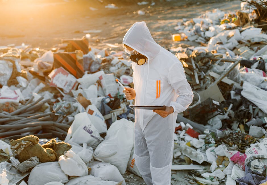 A person wearing a white protective suit with orange stripes, a full-face respirator mask, and gloves stands amidst a large, chaotic pile of mixed waste materials on a beach at sunset. The waste includes white plastic bags, foam insulation, broken pieces of drywall, and scattered debris of various textures and colors, including brown, gray, and black items. The individual is holding a clipboard in one hand and a handheld device in the other, appearing to inspect or survey the rubbish collection area. The environment is an outdoor coastal setting with the sunlight casting a warm glow over the scene, emphasizing the disorderly accumulation of waste. This image illustrates the context of independent waste management efforts, potentially involving hazardous or non-standard rubbish removal outside municipal services, highlighting the importance of proper disposal and environmental awareness in waste removal services.