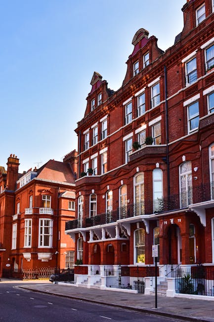 A multi-storey red brick residential building situated along a city street, featuring ornate architectural details such as tall, arched windows with white trim, decorative iron railings on the balconies, and a prominent curved roofline with gabled dormer windows. The building's façade displays a combination of textured brickwork and smooth stone accents, and its entrance is accessed via white-painted steps with black railings. The surrounding environment includes a pavement curb, a street sign, and parked vehicles, typical of an urban setting. The sky above is clear and bright, casting natural light that highlights the building's historic architecture and rich red tones. The scene reflects a residential area where private property owners might consider independent waste collection or rubbish removal services from providers like Waste Removal Kensington, especially for maintaining cleanliness without reliance on local authority services.