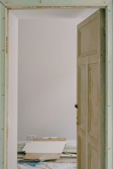 The image shows an interior view of a room under renovation, captured through an open wooden door with a natural finish featuring horizontal panels. The door is slightly ajar, revealing a white wall with no decorations or furnishings. In the lower part of the room, there are loose construction materials, including a white rectangular panel leaning against the wall, with some wooden planks and debris scattered on the floor, which is partly covered by protective sheeting or dust sheets. The environment appears to be a work-in-progress, with visible gaps in the door frame indicating ongoing installation or repairs. A double electrical socket is located near the floor, and the room is well-lit with natural light, giving a clear view of the plain, freshly painted wall and the unfinished state of the surrounding trim and doorframe. Waste removal specialists such as Waste Removal Kensington might be involved in clearing debris or coordinating private disposal of renovation waste in this setting, providing alternative waste handling options outside local authority collection methods.