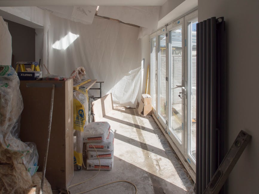 A construction or renovation project inside a room shows a partially finished space with some construction materials and clutter. On the left side, there are several large plastic bags filled with debris, which appear to be reusable or waste materials; some are tied at the top, and others are partially open, revealing insulation or packaging. Near the bags, a wooden or laminate countertop or work surface is visible, cluttered with various items, including a yellow level, a plastic container, and small tools. In the middle ground, a stack of white bags, likely containing cement or similar building materials, is placed on the unfinished concrete floor, which has visible dust and debris. To the right, a large black vertical radiator is mounted against the wall, adjacent to a set of large glass sliding doors that lead outside, allowing bright natural light to flood into the space. The doors feature a metal frame, and a wooden or metal threshold marks the boundary between the interior and exterior. Outside, a wooden fence can be seen through the glass. The room's ceiling is unfinished, with exposed plasterboard and insulation, and a white covering or curtain is draped near the light source. This scene suggests ongoing renovation work where Waste Removal Kensington could assist with private waste disposal or the removal of construction debris, supporting alternative waste handling methods during refurbishment processes.