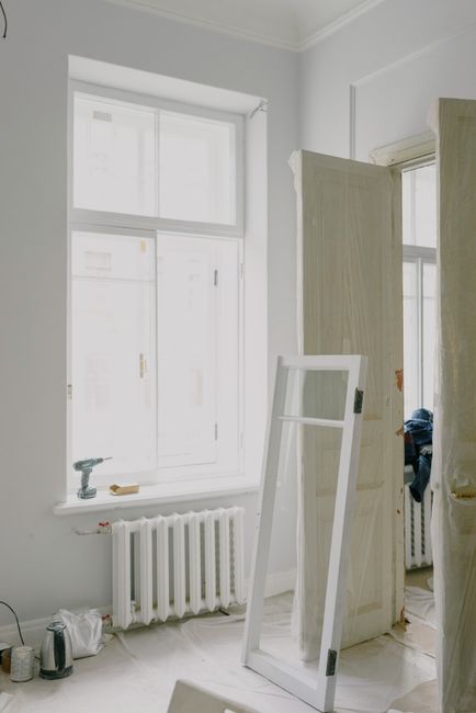 The image shows an interior view of a room under renovation, captured through an open wooden door with a natural finish featuring horizontal panels. The door is slightly ajar, revealing a white wall with no decorations or furnishings. In the lower part of the room, there are loose construction materials, including a white rectangular panel leaning against the wall, with some wooden planks and debris scattered on the floor, which is partly covered by protective sheeting or dust sheets. The environment appears to be a work-in-progress, with visible gaps in the door frame indicating ongoing installation or repairs. A double electrical socket is located near the floor, and the room is well-lit with natural light, giving a clear view of the plain, freshly painted wall and the unfinished state of the surrounding trim and doorframe. Waste removal specialists such as Waste Removal Kensington might be involved in clearing debris or coordinating private disposal of renovation waste in this setting, providing alternative waste handling options outside local authority collection methods.