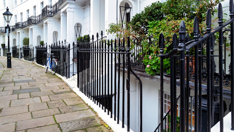 A row of white terraced houses with black wrought iron fences and railings along a paved sidewalk in a residential area. The fences feature pointed finials at the tops and are positioned on low white walls, with some sections enclosing small front gardens containing green foliage and flowering plants. On the right side, a bush with large green leaves and pink flowers partially obscures the fencing. A blue bicycle is leaning against the fence closer to the background. The scene is well-lit with natural daylight, and the sidewalk is composed of large, rectangular stone slabs with visible joints, showing some moss or dirt in the crevices. The overall environment suggests a typical urban or suburban street, where private property is delineated by decorative fencing, aligning with areas where waste removal or on-site clearance might be relevant within the context of independent waste disposal methods. The image emphasizes the neat, maintained appearance of the residential frontage, with careful attention to architectural details and greenery.
