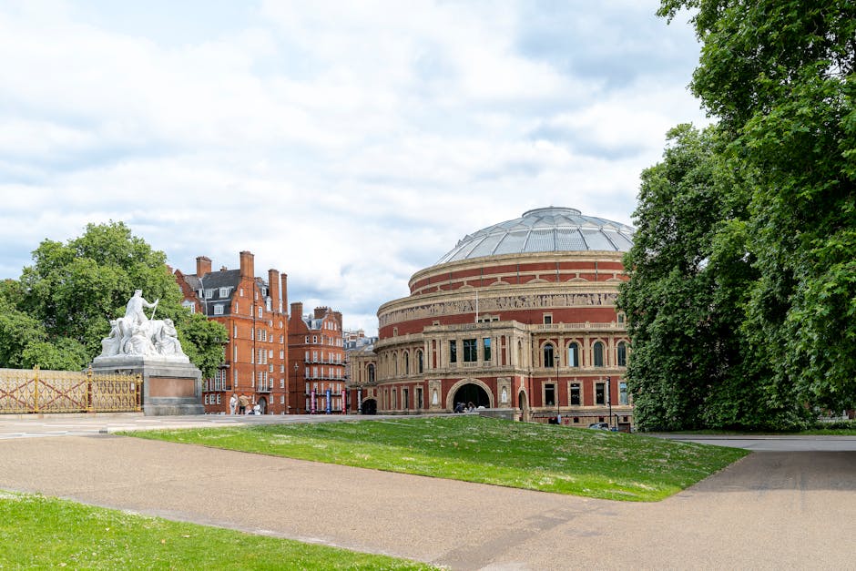 A wide view of a classical-style concert hall with a rounded brick facade and large arched entrance in an urban park setting. The building features decorative brickwork, tall narrow windows, and a domed roof with a metallic finish. Surrounding the hall are lush, green trees and a well-maintained lawn with a gravel pathway in the foreground, leading towards the structure. To the left, there is a white stone sculpture of a seated figure with a lion, placed on a podium, partially obscured by nearby trees. The scene is illuminated by soft, natural daylight under a partly cloudy sky, creating a calm and orderly atmosphere suitable for outdoor cultural events or casual visits. This scene may be relevant to private or alternative waste handling solutions such as rubbish collection services operating near public cultural landmarks, exemplifying how waste removal companies like Waste Removal Kensington support cleanliness in prominent city locations.