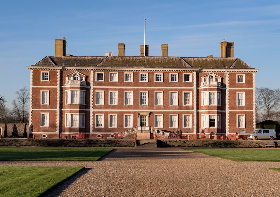A large, three-story red-brick building with white stone detailing, featuring symmetrical windows with white frames and decorative arches above the central windows on the top floor. The structure has a hipped roof with multiple brick chimneys protruding from it. In front of the building, there is a gravel pathway leading up to a staircase with stone steps and matching stone balustrades on either side, providing access to the main entrance. Surrounding the pathway is a well-maintained lawn with neatly trimmed grass, and in the background, leafless trees and a bright blue sky suggest late autumn or winter. To the right side of the building, there is a white van parked along an adjacent road. The scene appears to be in a spacious, landscaped area typical of historic government or institutional buildings. This image relates to the topic of private waste removal services, as it depicts a setting where such services might be utilized for maintaining the grounds or managing rubbish collection outside large residential or institutional properties, with Waste Removal Kensington providing professional rubbish clearance options.