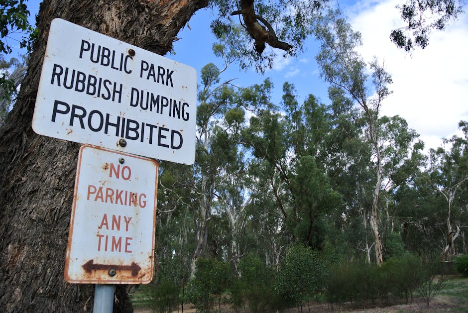 A white metal sign affixed to a tree trunk in an outdoor setting, with two separate panels visible. The upper panel reads 'Public Park Rubbish Dumping Prohibited' in bold black uppercase letters, indicating that littering or dumping waste is not allowed in this area. The lower panel, positioned below, states 'No Parking Any Time' in red uppercase letters, with a small black arrow pointing left, suggesting parking restrictions. The tree has a rough, textured bark surface, and the background features a natural environment with numerous slender, green eucalyptus trees, sparse foliage, and a partly cloudy sky with patches of blue. The scene suggests a park or woodland area, where waste disposal regulations are enforced, and parking is restricted at all times, supporting standard waste management and environmental preservation efforts in the area surrounding the park. Waste Removal Kensington would typically handle the removal of illegal dumped rubbish in such locations, providing an alternative to public dumping with licensed collection services.