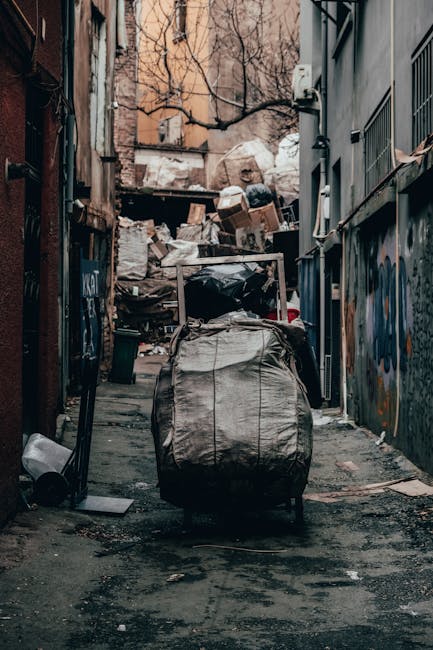 A narrow alleyway in an urban setting is filled with a large, black, and white flexible bag full of waste, positioned centrally on the damp, uneven asphalt ground. Behind the bag, a pile of various discarded cardboard boxes, plastic bags, and other rubbish clutter the background, stacked haphazardly against the wall of a building with peeling paint and graffiti. To the left, a small black trash bin and a white object, possibly a discarded piece of furniture or container, are visible on the ground. The surrounding environment includes brick and concrete structures, with metal security grilles over windows on the right side and exposed pipes attached to the building facades. Bare tree branches extend over the scene from the background, indicating an outdoor setting during a colder season. The lighting is natural, slightly muted, casting soft shadows, and the entire scene suggests informal or unmanaged waste accumulation, which Waste Removal Kensington could assist with through private rubbish collection and on-site clearance services.