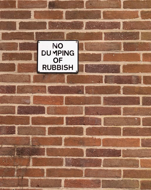 A white metal sign affixed to a tree trunk in an outdoor setting, with two separate panels visible. The upper panel reads 'Public Park Rubbish Dumping Prohibited' in bold black uppercase letters, indicating that littering or dumping waste is not allowed in this area. The lower panel, positioned below, states 'No Parking Any Time' in red uppercase letters, with a small black arrow pointing left, suggesting parking restrictions. The tree has a rough, textured bark surface, and the background features a natural environment with numerous slender, green eucalyptus trees, sparse foliage, and a partly cloudy sky with patches of blue. The scene suggests a park or woodland area, where waste disposal regulations are enforced, and parking is restricted at all times, supporting standard waste management and environmental preservation efforts in the area surrounding the park. Waste Removal Kensington would typically handle the removal of illegal dumped rubbish in such locations, providing an alternative to public dumping with licensed collection services.