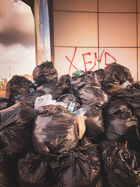A large pile of black plastic rubbish bags, some with visible stretching and creases, stacked against a tiled wall with beige and light brown squares. The bags contain various discarded items, including clear plastic water bottles, some partially crushed or crumpled, and bits of paper or packaging protruding from the openings. The backdrop features a metallic cylindrical structure partially visible on the left side, reflecting light, and a graffiti tag in red spray paint reading 'XEND' on the wall behind the bags. The scene is outdoors, with overcast sky visible in the upper left corner, casting diffuse lighting over the area. The surrounding environment suggests an urban setting, possibly near a commercial or residential building. The image relates to waste management and private rubbish collection services often provided by companies like Waste Removal Kensington, who handle on-site clearance and alternative disposal for such bulky refuse.