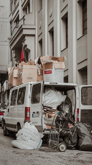 A white commercial van parked on a city street next to a multi-storey building with light-colored stone or concrete façade. The van's rear doors are open, revealing an interior filled with large white plastic bags, cardboard boxes, and miscellaneous waste materials. Several cardboard boxes, some labeled with red and black text, are stacked on top of the van, with others leaning against it, indicating the vehicle is being used for private rubbish collection or on-site clearance. On the pavement beside the van, there are additional black and white plastic bags filled with waste, along with a small hand trolley or dolly. The scene captures the urban environment typical for waste removal services provided by companies like Waste Removal Kensington, highlighting actions related to rubbish handling in a city setting, with natural, diffuse daylight illuminating the area, emphasizing the textures of the various waste materials and the urban architecture backdrop.