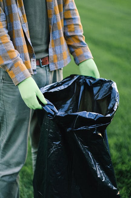 A person wearing a plaid shirt in shades of yellow, brown, and gray, along with green gloves, is holding open a large black rubbish bag made of shiny plastic material. The bag appears to be empty or in the process of being filled, with its top partially folded over. The individual is standing outdoors on a grass surface, which is lush and green, with a blurred background indicating a garden or park environment. The scene suggests an activity related to waste collection or rubbish removal, possibly part of an outdoor cleaning or yard clearing effort, which aligns with services offered by waste removal companies like Waste Removal Kensington. The natural daylight highlights the textures of the gloves, the bag, and the clothing, creating a clear, professional, and neutral depiction of waste management practices involving private or alternative disposal methods, distinct from municipal collection.