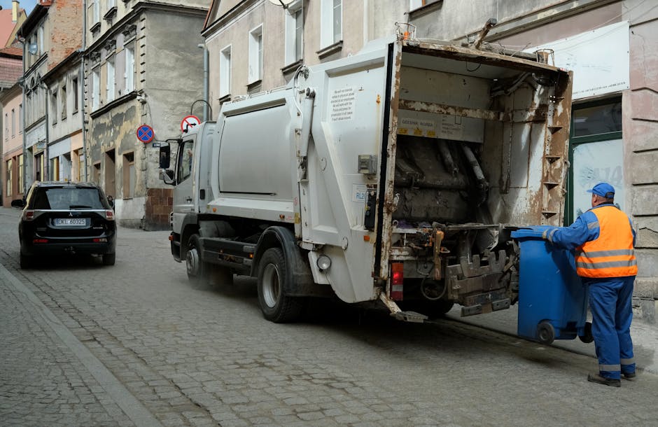 A waste collection vehicle operated by Waste Removal Kensington is parked on a narrow cobblestone street in an urban area. The truck is white with weathered, rusted metal components at the rear, which is open and exposing mechanisms for rubbish compaction. A worker dressed in a high-visibility orange vest and blue uniform is standing beside the truck, operating a blue wheeled rubbish bin prepared for collection. The street is lined with multi-storey residential buildings featuring painted and weathered facades, with some apartments having small windows and decorative elements. A black car is parked further down the street, and a no-parking sign is visible on the building frontage. The environment appears quiet and typical of an inner-city neighborhood, with natural daylight illuminating the scene, emphasizing the functional, utilitarian design of the waste collection process involved in private or alternative waste handling services around Kensington.