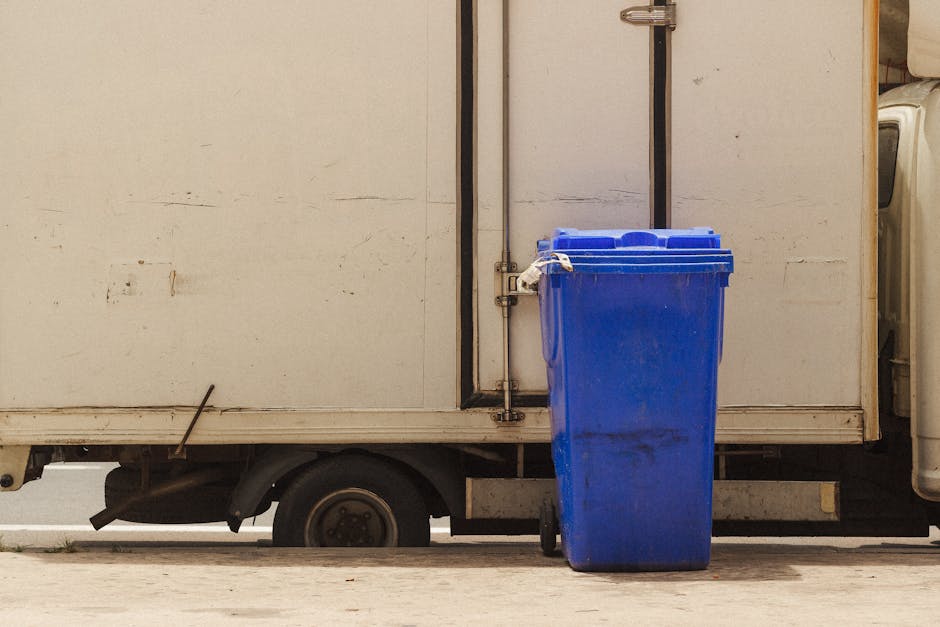 The image depicts a blue wheeled rubbish bin positioned on a paved surface directly beside a white delivery truck with a metal exterior, which features visible rust and dirt marks. The bin has a textured surface with slight grime stains near its bottom and a closed lid on top. It is placed to the right of the truck's rear door, which is secured with a metal latch and hinges. The truck's side panel appears weathered, with signs of wear such as small scratches and discoloration, and the rear wheel is partially visible beneath the cargo area. A vertical black metal pole runs along the back of the truck, supporting the door mechanism. The environment appears to be an outdoor urban or industrial area with natural daylight illuminating the scene. This setup suggests an on-site collection or private waste handling scenario, consistent with rubbish removal services, such as those offered by Waste Removal Kensington. The scene emphasizes the practical aspects of alternative waste disposal methods, typical in rubbish management contexts for bulky or scheduled waste pickups.